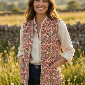 Puede incluir: Un chaleco con estampado floral con fondo crema y flores rojas. El chaleco tiene cierre de botones y bolsillos. La persona lleva una camisa blanca y vaqueros azules. El fondo es un campo de flores silvestres.