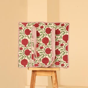 May include: A square tote bag with a repeating pattern of red pomegranates and green leaves on a cream background. The bag has two red handles and a red stitched border. The bag is sitting on a small wooden stool.