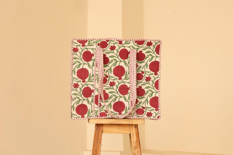 May include: A square tote bag with a repeating pattern of red pomegranates and green leaves on a cream background. The bag has two red handles and a red stitched border. The bag is sitting on a small wooden stool.