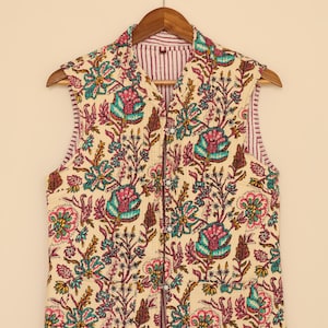 May include: Cream quilted vest featuring a floral design in teal, pink, and brown. The vest has a striped lining and a button-up front. It is displayed on a wooden hanger.