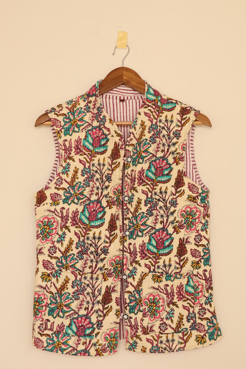 May include: Cream quilted vest featuring a floral design in teal, pink, and brown. The vest has a striped lining and a button-up front. It is displayed on a wooden hanger.