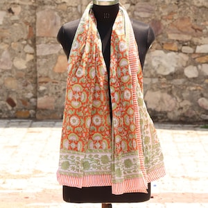 May include: A vibrant orange scarf with a floral pattern in white and green, displayed on a mannequin. The scarf has a striped border in red and white. The background is a textured stone wall.