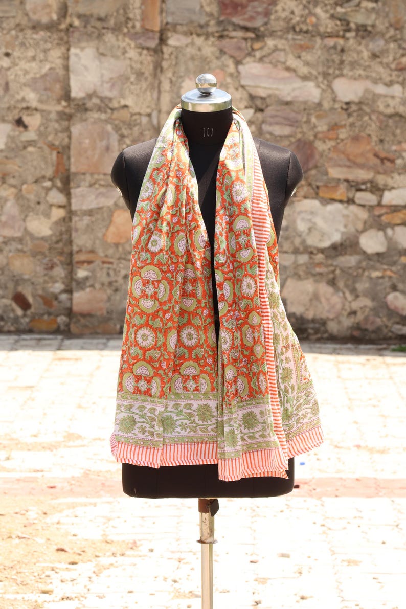 May include: A vibrant orange scarf with a floral pattern in white and green, displayed on a mannequin. The scarf has a striped border in red and white. The background is a textured stone wall.