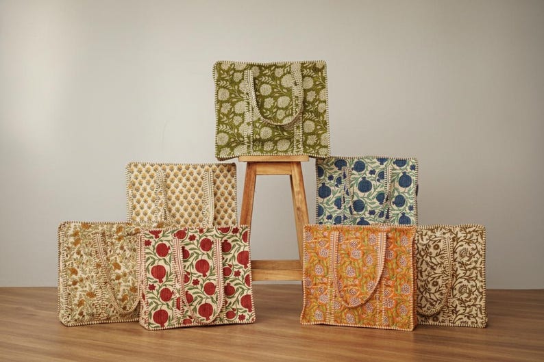 May include: A collection of tote bags with floral patterns in various colors, including green, red, blue, and orange. The bags are made of fabric and have matching handles. Some bags are stacked on a wooden stool.