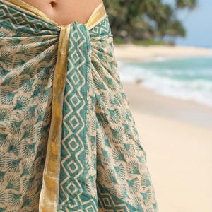 Könnte beinhalten: Ein beigefarbenes und türkisfarbenes Sarong mit einem geometrischen und floralen Muster, das um die Taille gebunden ist. Das Sarong hat einen goldenen Rand und vor einem Strandhintergrund mit Palmen und Wellen.