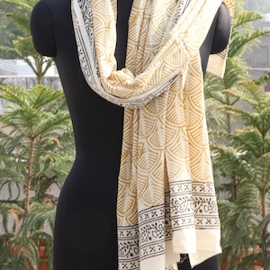 Pode incluir: Len&ccedil;o creme com um padr&atilde;o repetitivo dourado em forma de leque e bordas pretas, drapeado sobre um manequim preto. O len&ccedil;o &eacute; feito de tecido leve. O fundo apresenta vegeta&ccedil;&atilde;o. O comprimento do len&ccedil;o &eacute; de aproximadamente 180 cm.