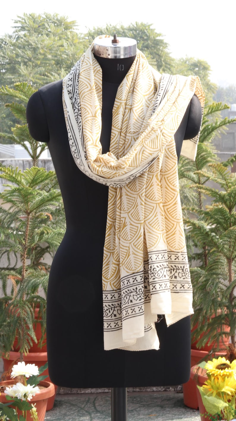 Pode incluir: Len&ccedil;o creme com um padr&atilde;o repetitivo dourado em forma de leque e bordas pretas, drapeado sobre um manequim preto. O len&ccedil;o &eacute; feito de tecido leve. O fundo apresenta vegeta&ccedil;&atilde;o. O comprimento do len&ccedil;o &eacute; de aproximadamente 180 cm.