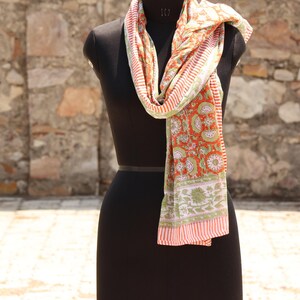 May include: A colorful scarf with a floral pattern in shades of orange, green, and white is draped over a black mannequin. The scarf has a striped border and a detailed design.
