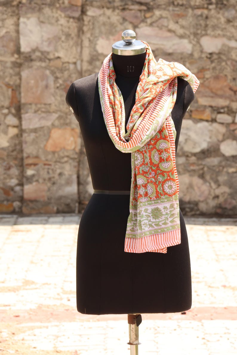 May include: A colorful scarf with a floral pattern in shades of orange, green, and white is draped over a black mannequin. The scarf has a striped border and a detailed design.