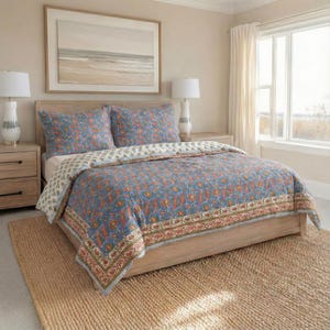 May include: A bedroom scene featuring a bed with a blue floral patterned quilt and matching pillowcases. The quilt has a red and green border. The bed sits on a light wood frame, with a woven rug on the floor. Two lamps and a framed picture are also visible.