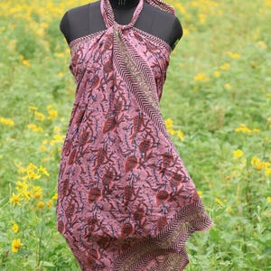 May include: A pink sarong with a floral pattern, displayed on a mannequin. The fabric features a repeating design of leaves and flowers in shades of brown and blue. The edges are trimmed with a patterned border. The sarong is tied around the neck.