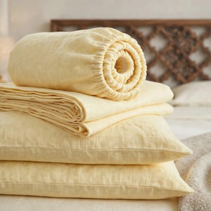 Peut inclure: Un ensemble de literie jaune pâle comprenant un drap-housse roulé en cylindre avec des bords élastiques visibles, un drap plat plié soigneusement et deux taies d'oreiller rectangulaires empilées. Le tissu présente une texture tissée.