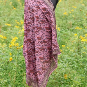 May include: A pink scarf with a floral pattern and a geometric border is draped over a mannequin. The scarf features a repeating design of brown and navy blue floral elements on a pink background. The border has a chevron pattern.