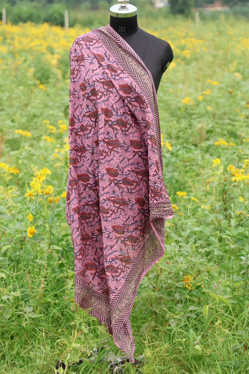May include: A pink scarf with a floral pattern and a geometric border is draped over a mannequin. The scarf features a repeating design of brown and navy blue floral elements on a pink background. The border has a chevron pattern.