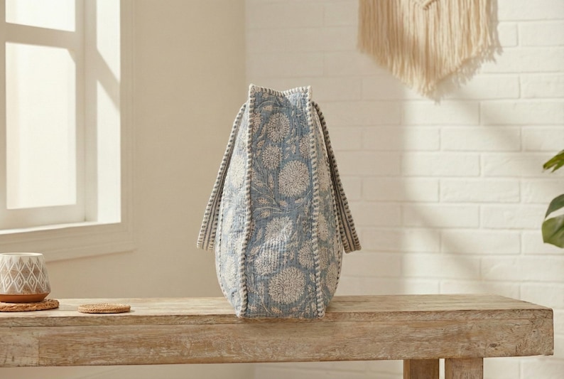 May include: A blue and white floral patterned tote bag with two handles. The bag is made of a textured fabric and sits on a wooden bench. The bag's design features a repeating floral pattern.