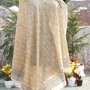 Pode incluir: Um xale creme com um padr&atilde;o repetitivo em forma de leque em um tom amarelo dourado. O xale tem um design de borda em preto e branco. O tecido &eacute; drapeado sobre um manequim, mostrando o design e a textura.