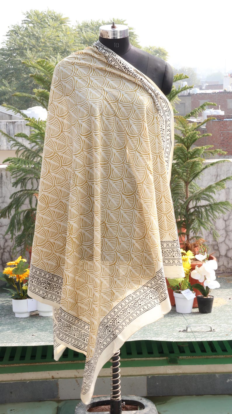 Pode incluir: Um xale creme com um padr&atilde;o repetitivo em forma de leque em um tom amarelo dourado. O xale tem um design de borda em preto e branco. O tecido &eacute; drapeado sobre um manequim, mostrando o design e a textura.