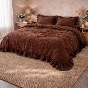 May include: A bedroom scene featuring a bed with a dark brown ruffled duvet cover and matching pillowcases. The bed sits on a woven jute rug. A wooden nightstand with a lamp and flowers is visible.