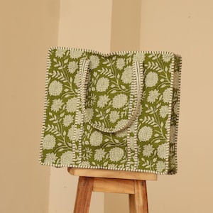 May include: A green tote bag with a floral pattern in white. The bag has a rectangular shape with a handle and is sitting on a wooden stool. The bag's edges and handle are trimmed with a white border.