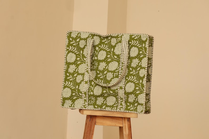 May include: A green tote bag with a floral pattern in white. The bag has a rectangular shape with a handle and is sitting on a wooden stool. The bag's edges and handle are trimmed with a white border.