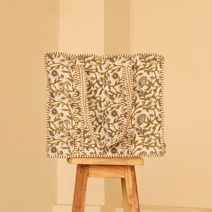 May include: A square tote bag with a floral pattern in olive green and gray on an off-white background. The bag has two handles and is sitting on a small wooden stool. The edges of the bag are trimmed with a brown stitch.