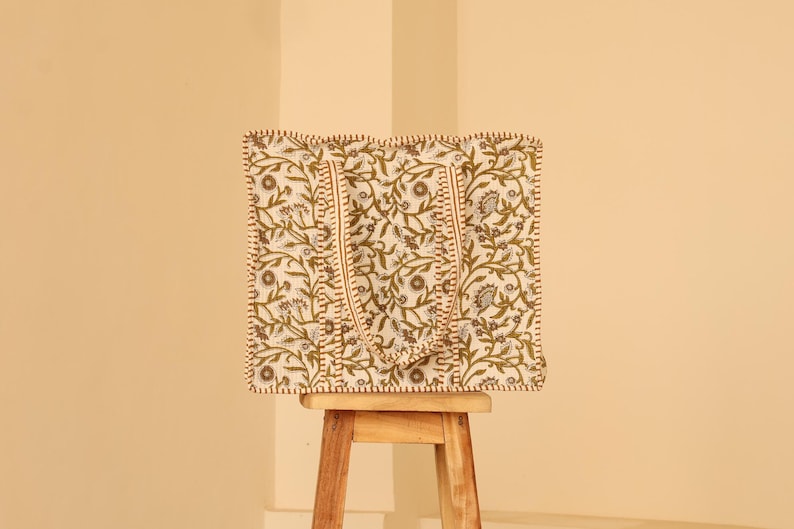 May include: A square tote bag with a floral pattern in olive green and gray on an off-white background. The bag has two handles and is sitting on a small wooden stool. The edges of the bag are trimmed with a brown stitch.