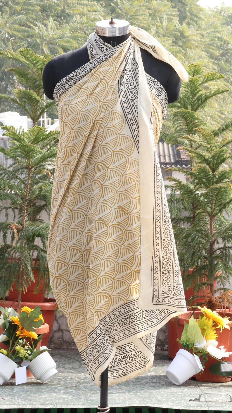 Pode incluir: Um envolt&oacute;rio t&ecirc;xtil creme com um padr&atilde;o de folhas repetido em dourado e detalhes de borda preta. O envolt&oacute;rio &eacute; drapeado sobre um manequim, mostrando seu design e textura. O fundo apresenta vegeta&ccedil;&atilde;o.