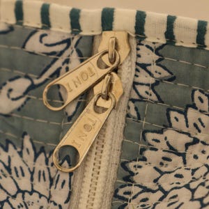 May include: Close-up of a section of a textile with a floral pattern in white and blue. Two gold-colored zippers are visible, with the word "TONI" engraved on each zipper pull. The fabric has a quilted appearance.