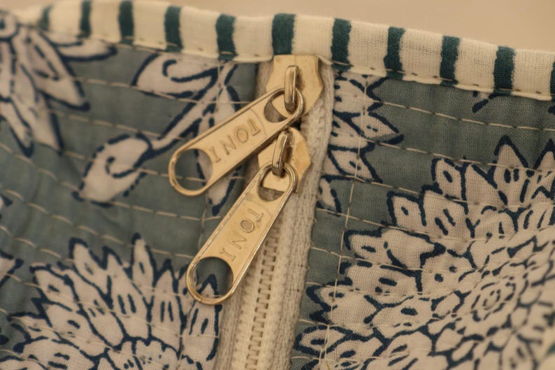 May include: Close-up of a section of a textile with a floral pattern in white and blue. Two gold-colored zippers are visible, with the word "TONI" engraved on each zipper pull. The fabric has a quilted appearance.