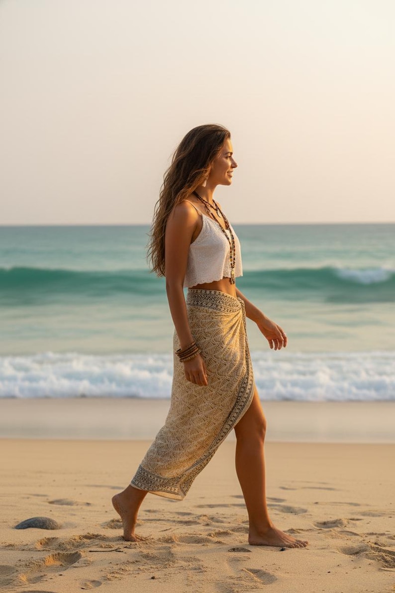 Pode incluir: Uma mulher com um top branco e um sarongue bege com um padr&atilde;o geom&eacute;trico caminha numa praia arenosa. Ela usa colares e pulseiras em camadas. O oceano e o c&eacute;u s&atilde;o vis&iacute;veis ao fundo.