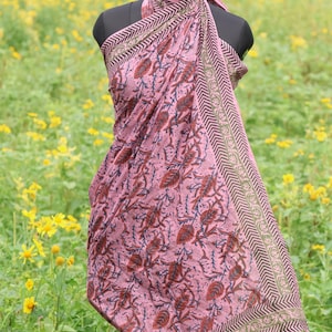 May include: A pink sarong with a repeating leaf pattern in brown and blue. The sarong is draped over a mannequin and has a decorative border with a geometric design in brown and green. The background is a field of yellow flowers.