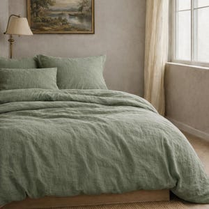 May include: A sage green linen bedding set on a wooden bed frame. The set includes a duvet cover and two pillowcases. A framed landscape painting hangs above the bed, and a beige rug is on the floor. A lamp and window with sheer curtains are also visible.
