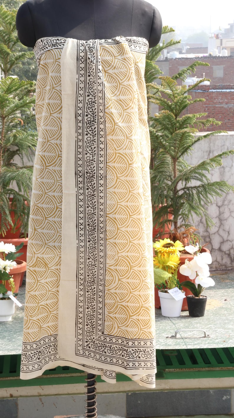 Pode incluir: Um tecido creme com um padr&atilde;o amarelo repetido e uma borda central em preto e branco. O tecido &eacute; drapeado sobre um manequim, mostrando o design e a textura. O tecido &eacute; provavelmente um len&ccedil;o ou xale.