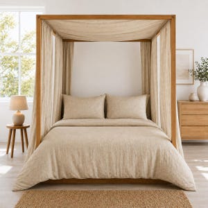 Op de afbeelding: Een hemelbedframe van lichtgekleurd hout, voorzien van een overkapping gedrapeerd met beige linnen stof. Het bed is opgemaakt met een bijpassend beige linnen dekbedovertrek en twee rechthoekige kussens voor een samenhangend geheel.
