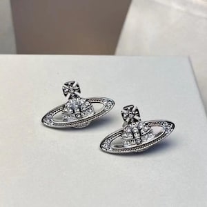 May include: A pair of silver-toned stud earrings. Each earring features a small orb design with a cross on top, encrusted with clear crystals. The earrings are displayed on a white box, with a white bag and a branded paper bag in the background.