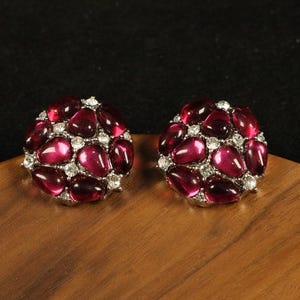 Könnte beinhalten: Ein Paar runde Ohrstecker. Jeder Ohrring hat eine Ansammlung von tiefroten, oval geformten Edelsteinen, die von kleineren, klaren Steinen umgeben sind. Die Ohrringe sind in einem silberfarbenen Metall gefasst und liegen auf einer Holzoberfläche.
