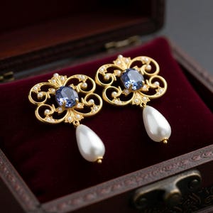 May include: A pair of ornate gold-colored earrings with a blue gemstone center and a dangling white teardrop pearl. The earrings are displayed in an open, dark wooden jewelry box with a red velvet interior.