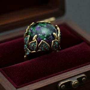 May include: A gemstone ring with a large, round, green and purple stone, set in a gold-colored metal. The ring is in a wooden box with a burgundy velvet interior. The ring's design includes leaf-like accents with small blue gemstones.