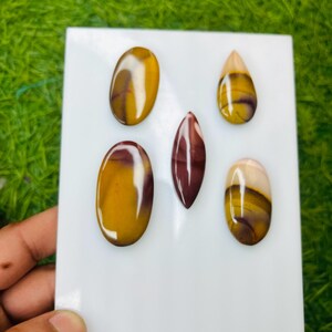 Può includere: Una collezione di sei pietre preziose lucide di varie forme, tra cui ovali e a goccia. Le pietre presentano un mix di colori, tra cui giallo, marrone e bordeaux, con una finitura lucida. Le pietre preziose sono esposte su una superficie bianca.