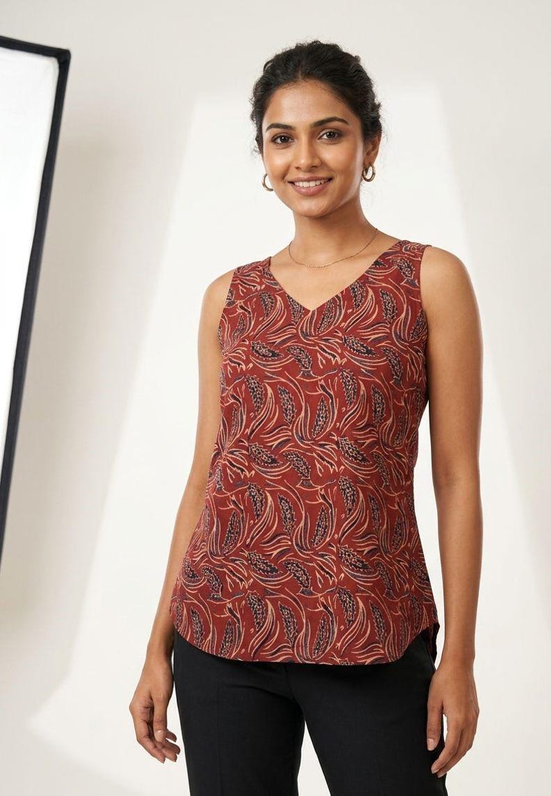 Puede incluir: Una blusa sin mangas con cuello en V, de color marr&oacute;n rojizo, adornada con un patr&oacute;n repetido de formas estilizadas en forma de hoja en tonos beige y marr&oacute;n oscuro. La blusa se combina con pantalones negros.