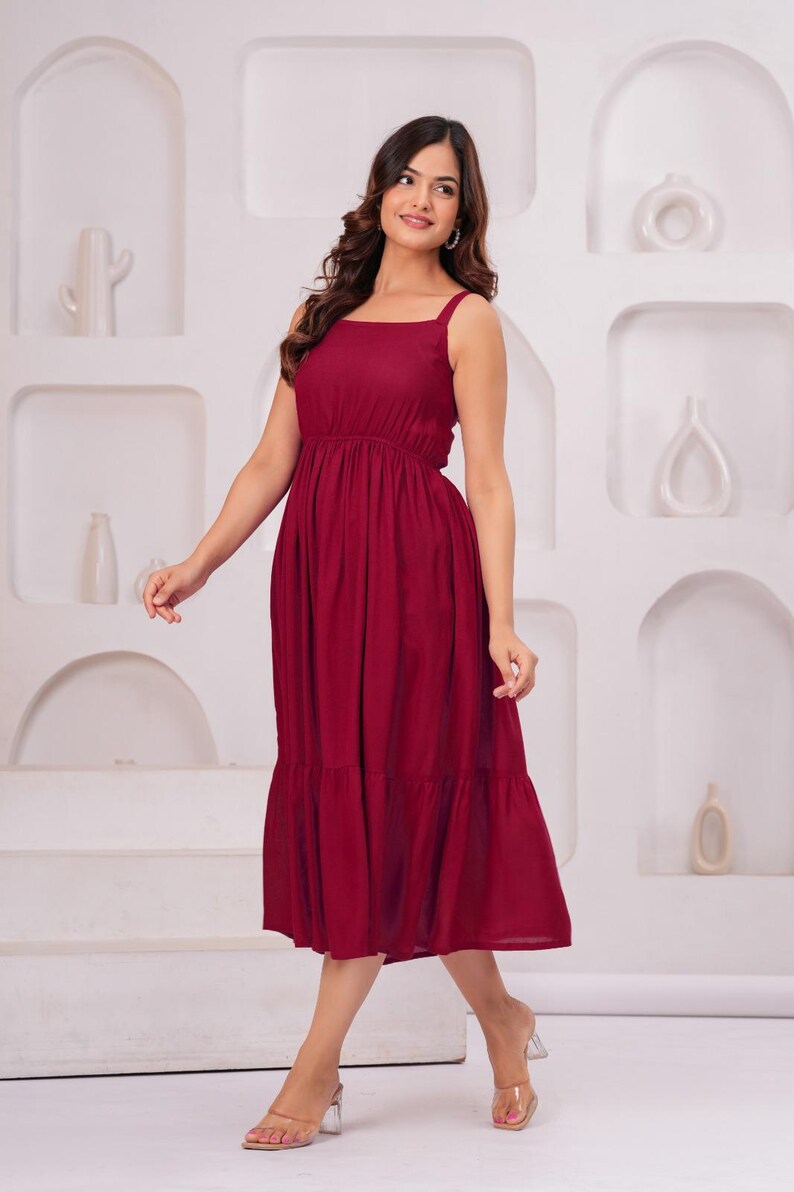 May include: A burgundy midi dress with a square neckline and thin straps. The dress features a fitted bodice, a gathered waist, and a tiered skirt. The model is wearing clear heeled sandals. The background is a white wall with decorative alcoves.