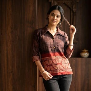 May include: A brown button-down shirt with a red patterned band at the waist and sleeves. The shirt has a collared neckline and three-quarter sleeves. The shirt is paired with black jeans.