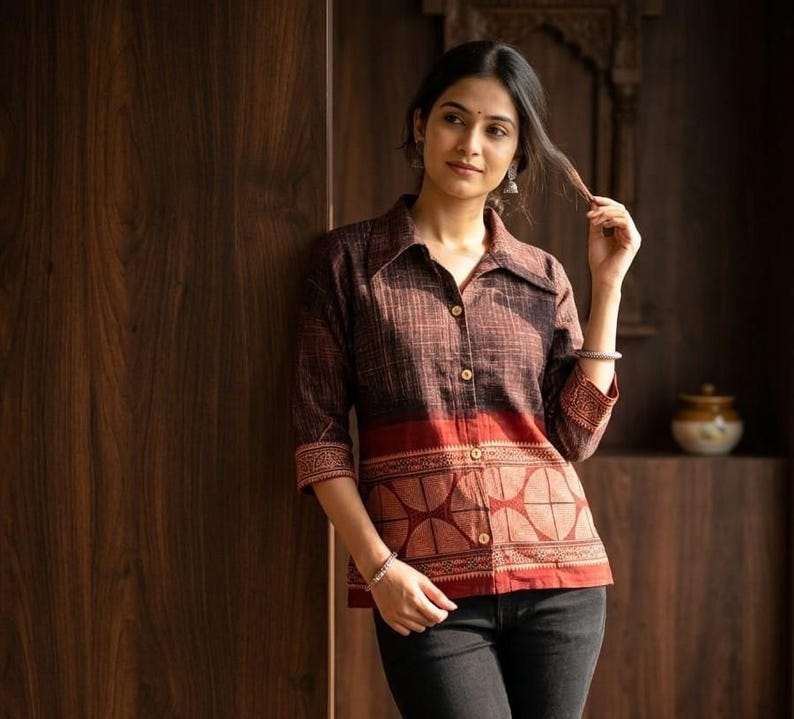 May include: A brown button-down shirt with a red patterned band at the waist and sleeves. The shirt has a collared neckline and three-quarter sleeves. The shirt is paired with black jeans.