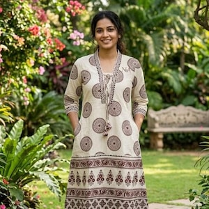 May include: A cream-colored dress with a brown circular pattern and decorative trim. The dress has long sleeves, pockets, and a V-neckline with a tassel tie. The dress is knee-length and worn outdoors in a garden setting.