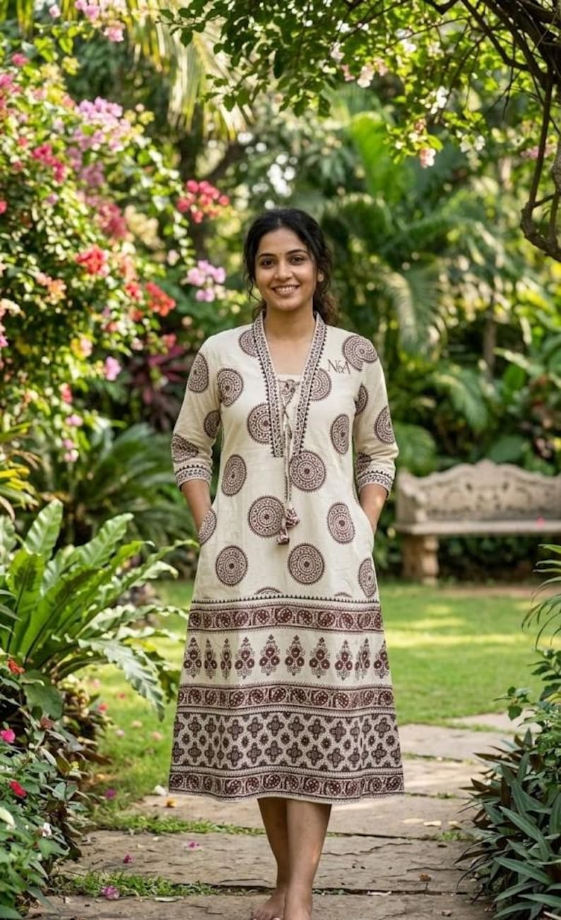 May include: A cream-colored dress with a brown circular pattern and decorative trim. The dress has long sleeves, pockets, and a V-neckline with a tassel tie. The dress is knee-length and worn outdoors in a garden setting.