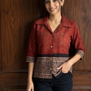 May include: A button-down shirt with a red and black patterned design. The shirt has three-quarter sleeves and a collared neckline. The bottom portion of the shirt features a black band with a detailed pattern. The model is wearing black jeans.
