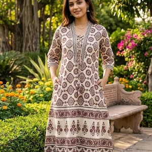 May include: A long, cream-colored dress with a brown floral pattern. The dress has a V-neck, long sleeves, and pockets. The model is standing in a garden setting with trees and flowers.