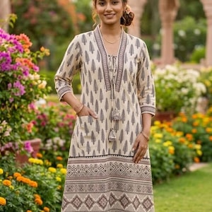 May include: A cream-colored dress with a black leaf pattern and geometric border. The dress has a V-neck, three-quarter sleeves, and a tassel tie. It features pockets and is knee-length. The woman is standing in a garden with colorful flowers.