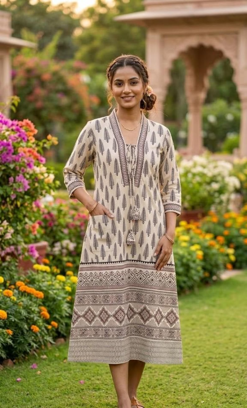 May include: A cream-colored dress with a black leaf pattern and geometric border. The dress has a V-neck, three-quarter sleeves, and a tassel tie. It features pockets and is knee-length. The woman is standing in a garden with colorful flowers.