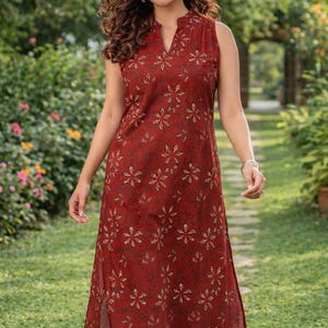 May include: A sleeveless, knee-length dress in a deep red hue, adorned with a repeating floral pattern in a lighter shade. The dress features a mandarin collar and side slits. The model is wearing white leggings.